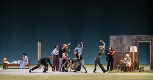 en stor gruppe dansere og skuespillere på scenen, en seng, lenestol og stålampe