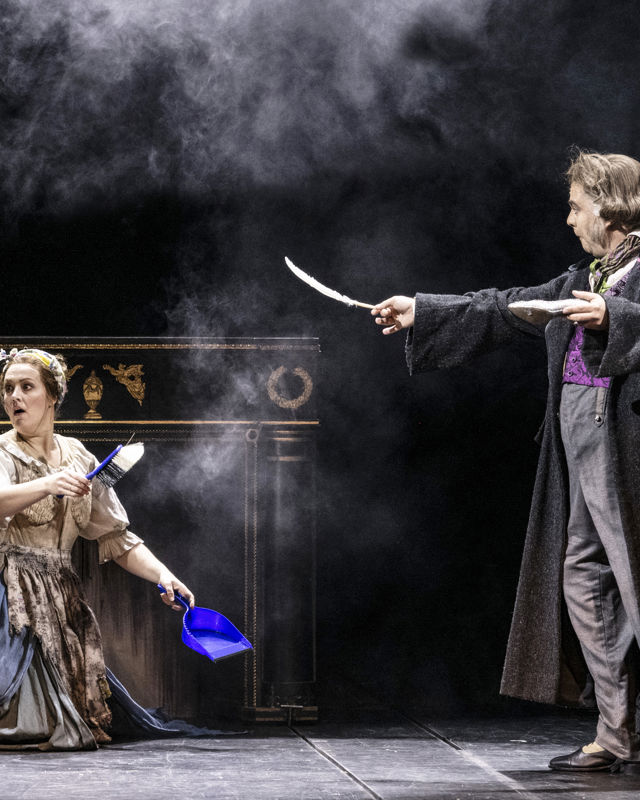 Kvinne sitter på kne foran kamin med askesky rundt, mann med frakk og englevinger ved siden av