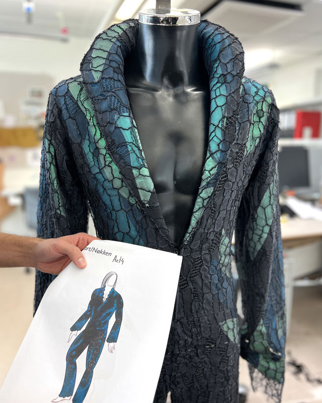Nøkkens kostyme i turkise farger med svart heklede snorer over. En hånd holder en kostymetegning foran.