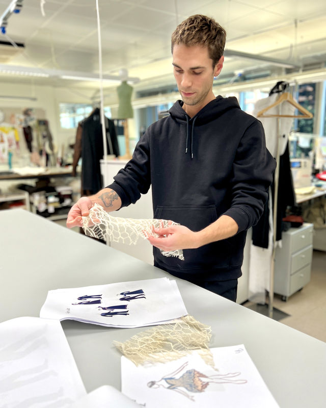 En mann holder fram et stykke lyst, hullet stoff over et bord med kostymetegninger av en ballettdanser i svaneaktig drakt og herrer i dress.