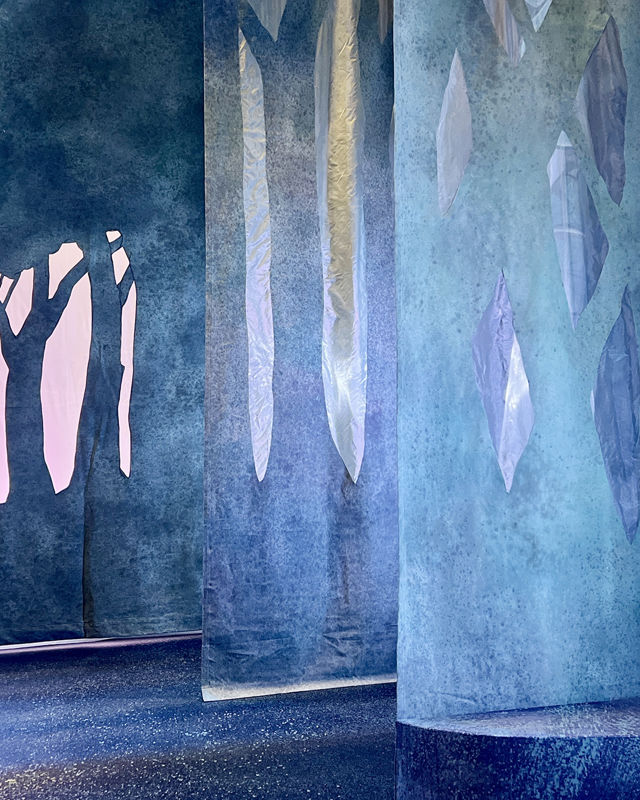 Blå-turkisfargede tepper viser lag på lag innover mot sceneteppet med hus og trær.