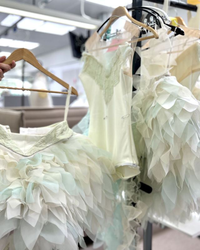 Fjærlignende svanekostymer i hvite toner ispedd litt turkis henger på stativ. To hender løfter fram ett av kostymene.