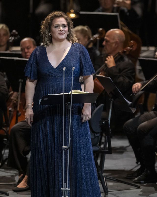 Kvinne i lang blå kjole foran orkestermusikere