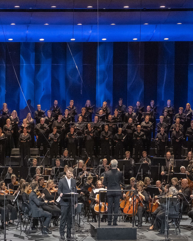 Mann i dress på scenen foran stort orkester og kor