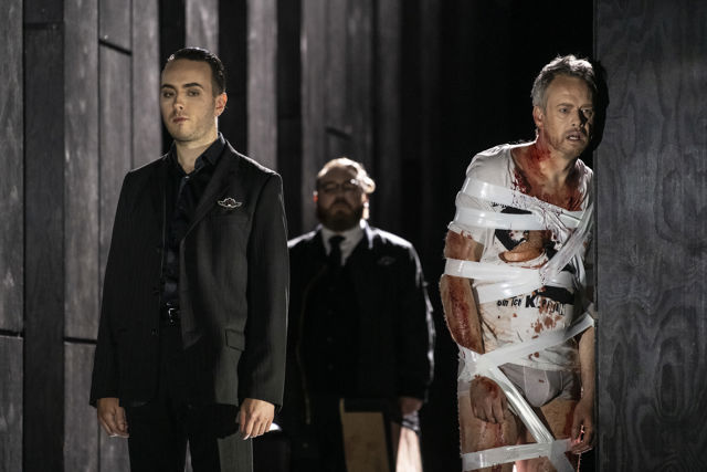 Blodig mann surret inn i tape, to menn i dress betrakter